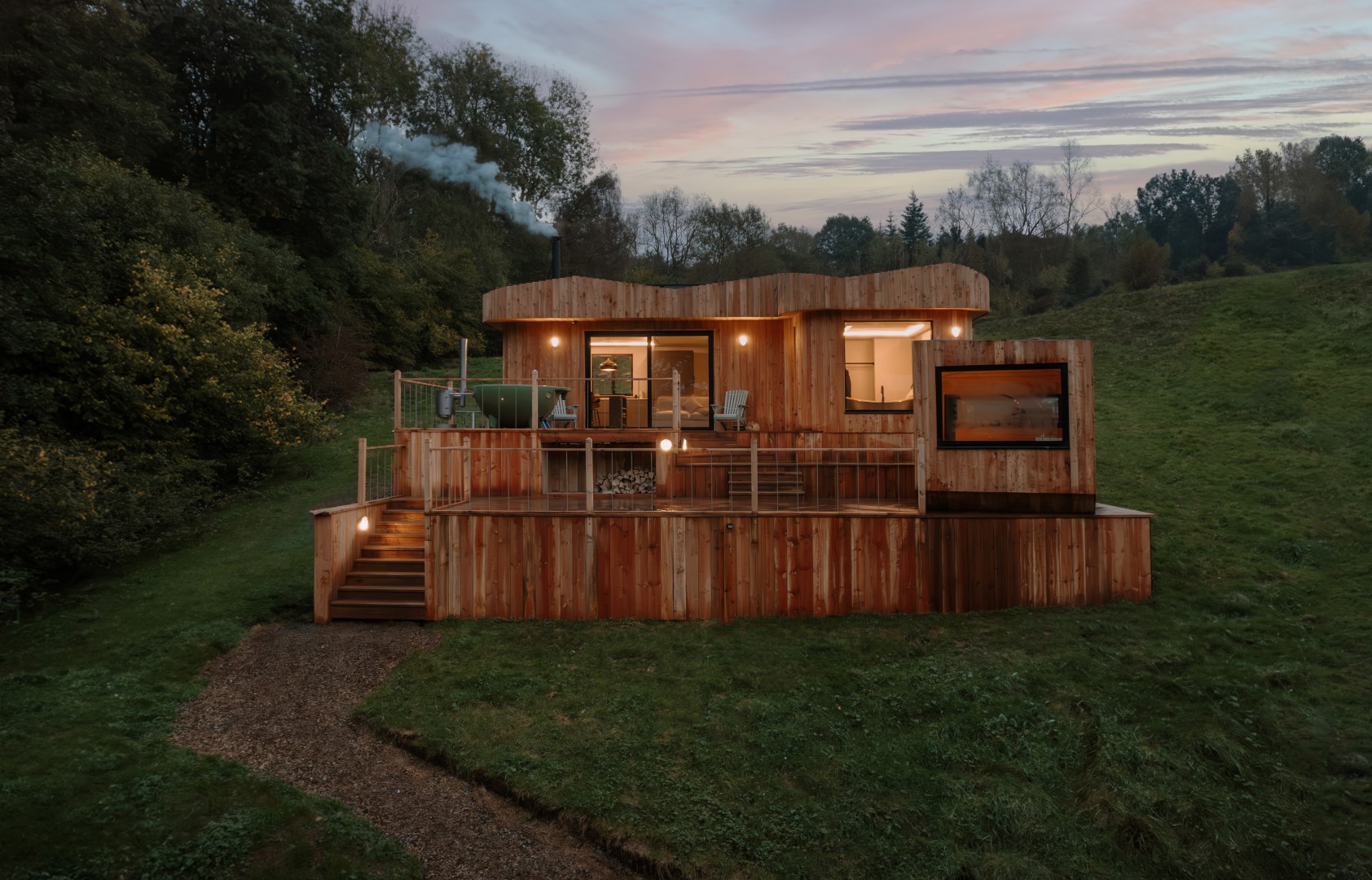 Scandi Cabin nestled on a grassy hill, featuring large windows and a deck perfect for sunset views, with trees forming a serene backdrop.