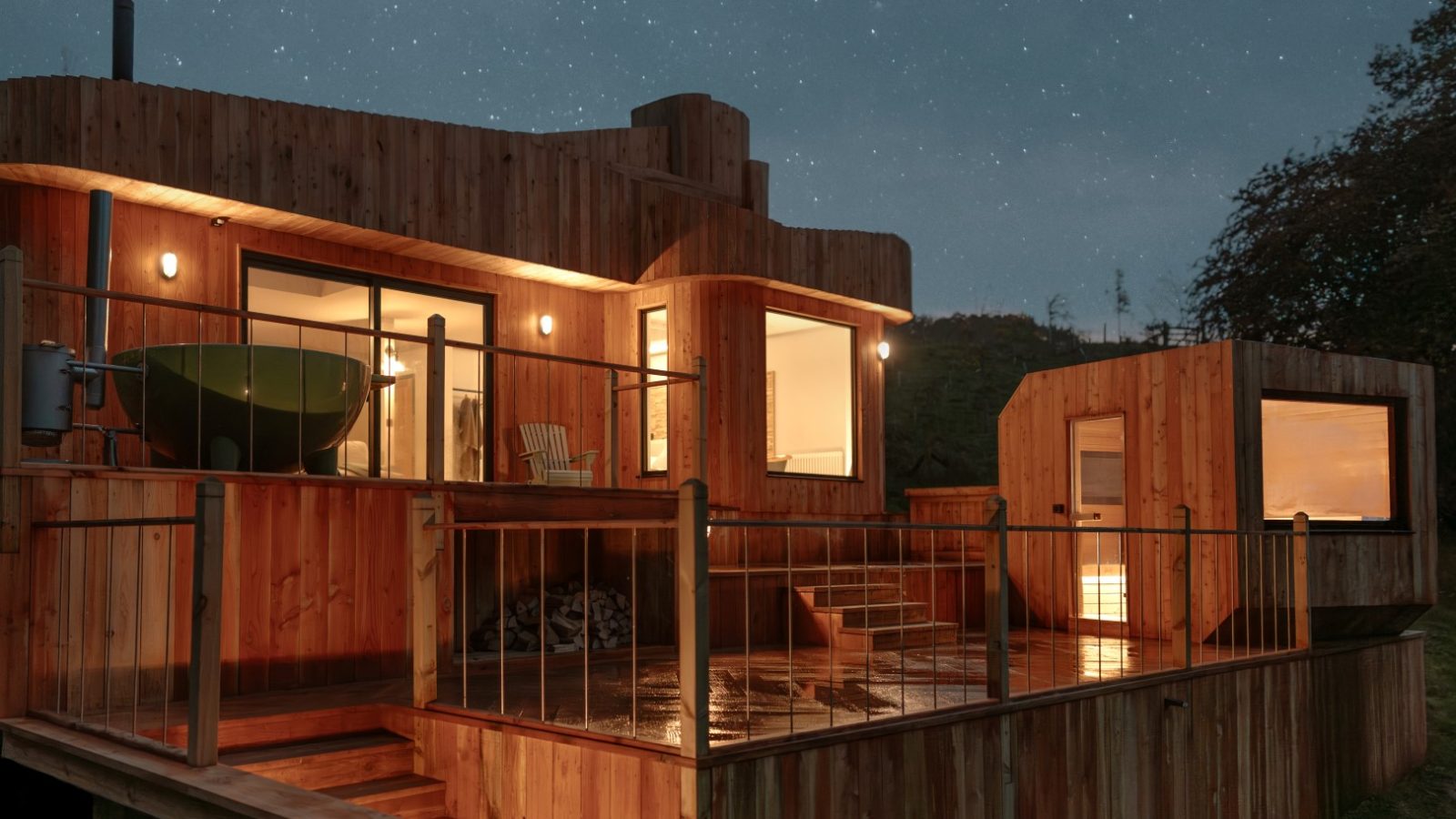 A modern Scandi Cabin with large windows, a deck, and outdoor lights, set against a starry night sky.