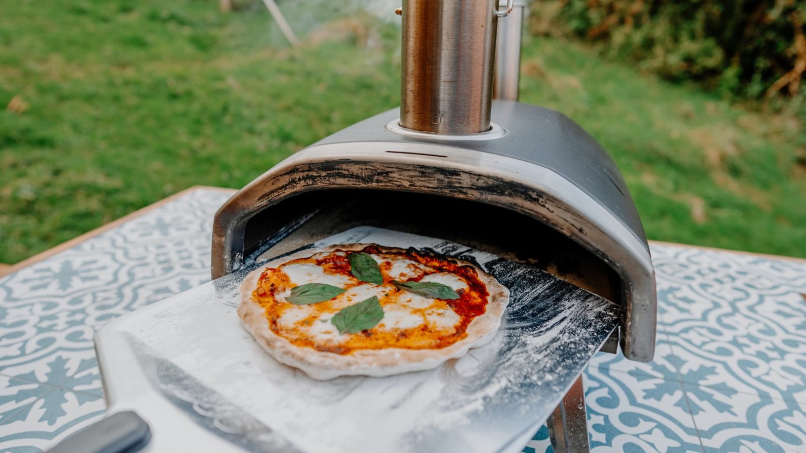 A pizza with fresh basil and bubbling cheese rests on a metal peel as it's pulled from The Fold's cozy outdoor pizza oven, set against a charming patterned surface reminiscent of a Scandi cabin retreat.