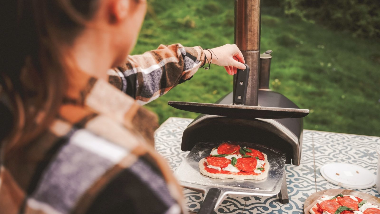 A person places a pizza with pepperoni and basil into an outdoor pizza oven on a patterned table at the picturesque Scandi Cabin at The Fold.