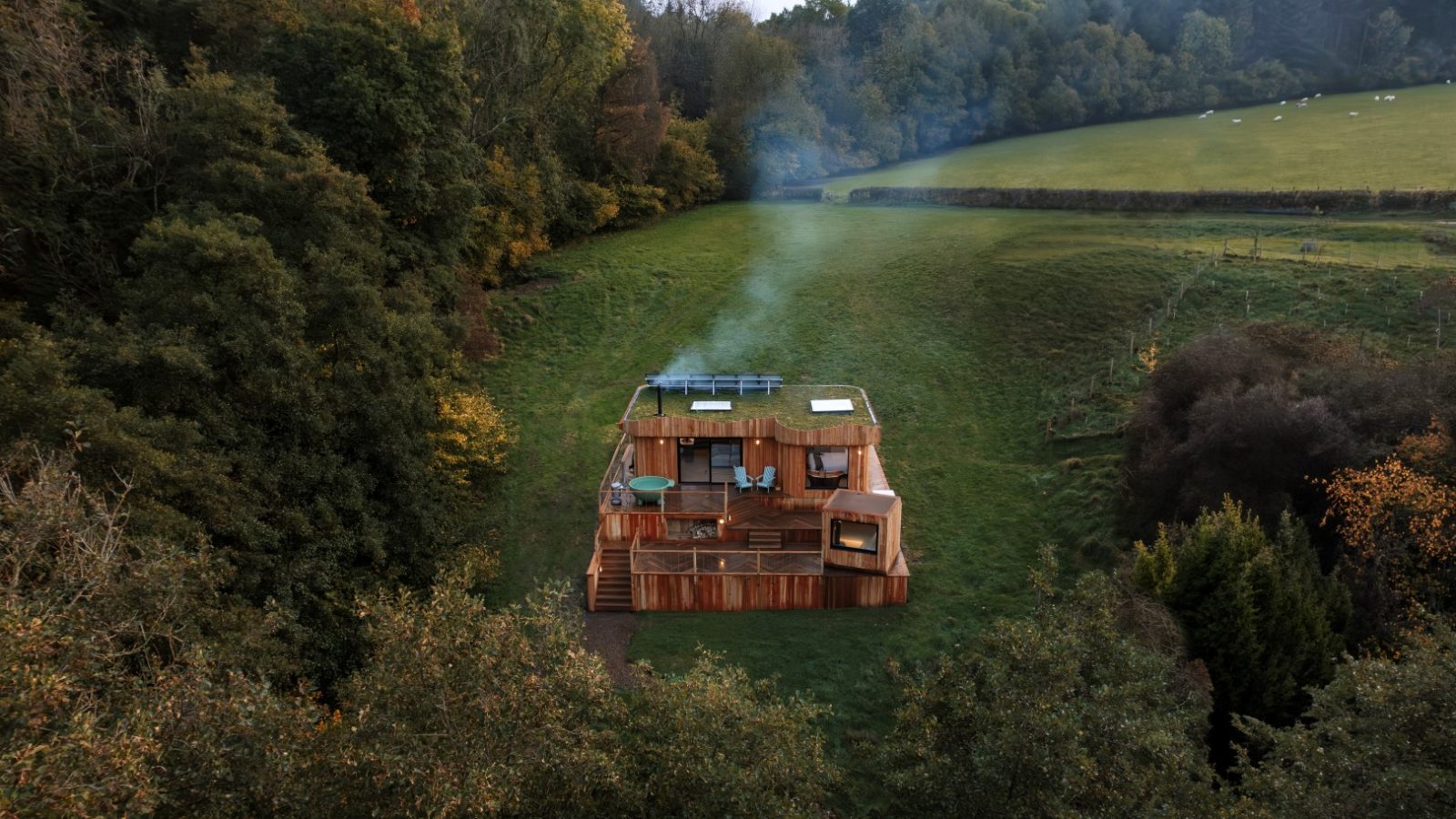 Aerial view of The Fold, a charming wooden cabin with a hot tub and outdoor seating, nestled among trees and an open grassy field.