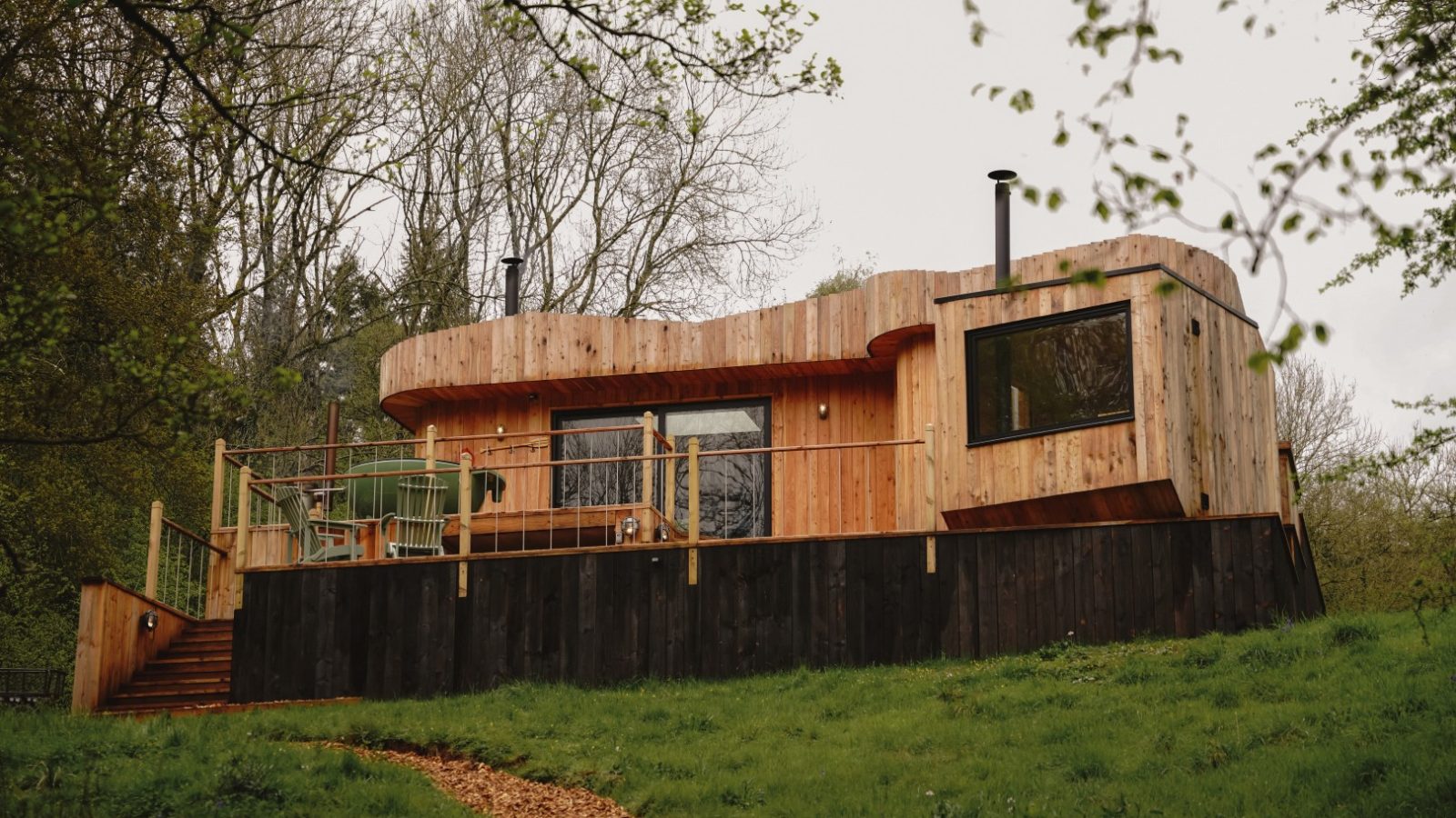 The Scandi Cabin boasts large windows and a deck, nestled amid trees in a lush grassy area.