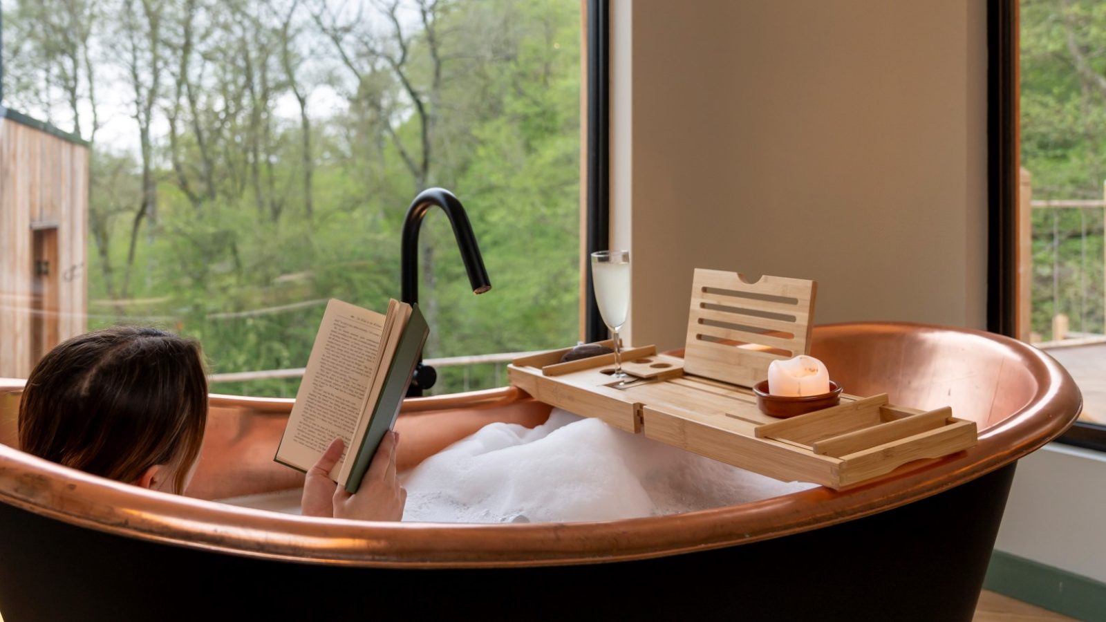 In a cozy Scandi cabin, a person relaxes in a copper bathtub, absorbed in a book. A wooden tray holds a flickering candle and a glass, nestled by the side of the tub near a window that offers serene views of The Fold's lush trees.