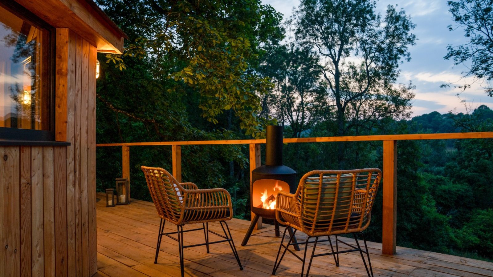 On the outdoor wooden deck of The Fold, two wicker chairs face a lit fireplace, surrounded by lush trees at dusk.