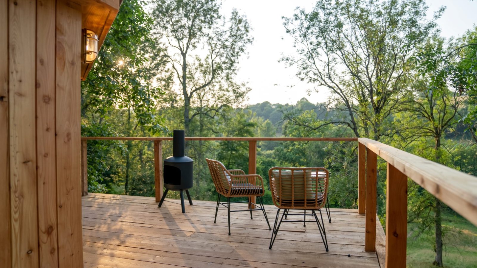 Perched high in the treehouse, this wooden deck features two wicker chairs and a small stove, offering a stunning view of The Fold's lush greens and clear skies.