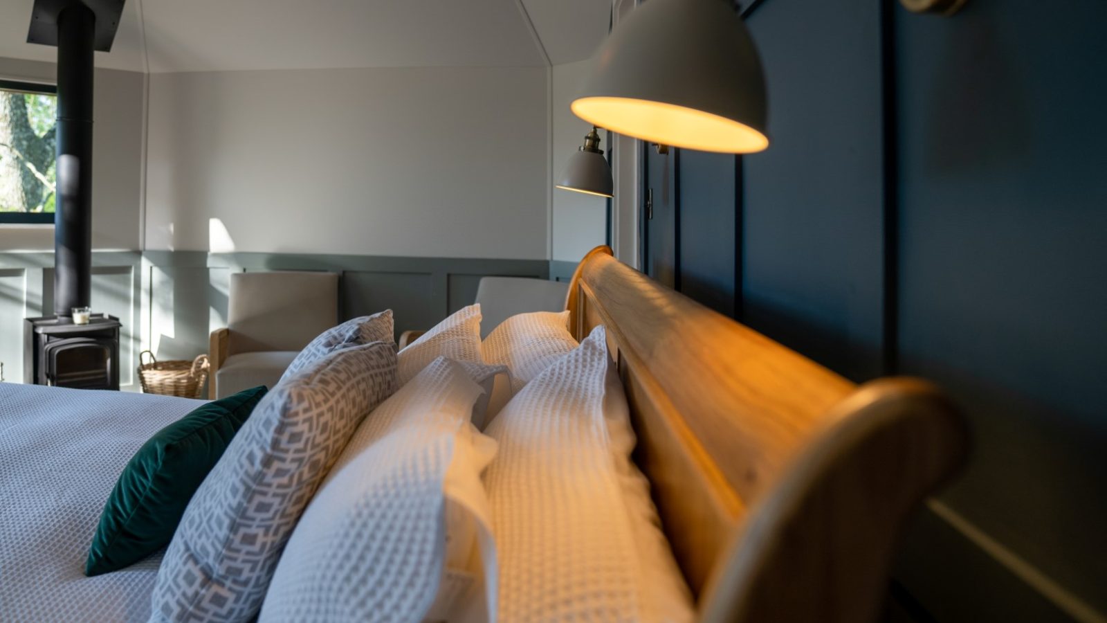 Cozy bedroom in The Fold, featuring a wooden sleigh bed adorned with white and patterned pillows beneath a gently glowing wall lamp. A treehouse-like charm is captured with a chair and wood stove nestled in the background.