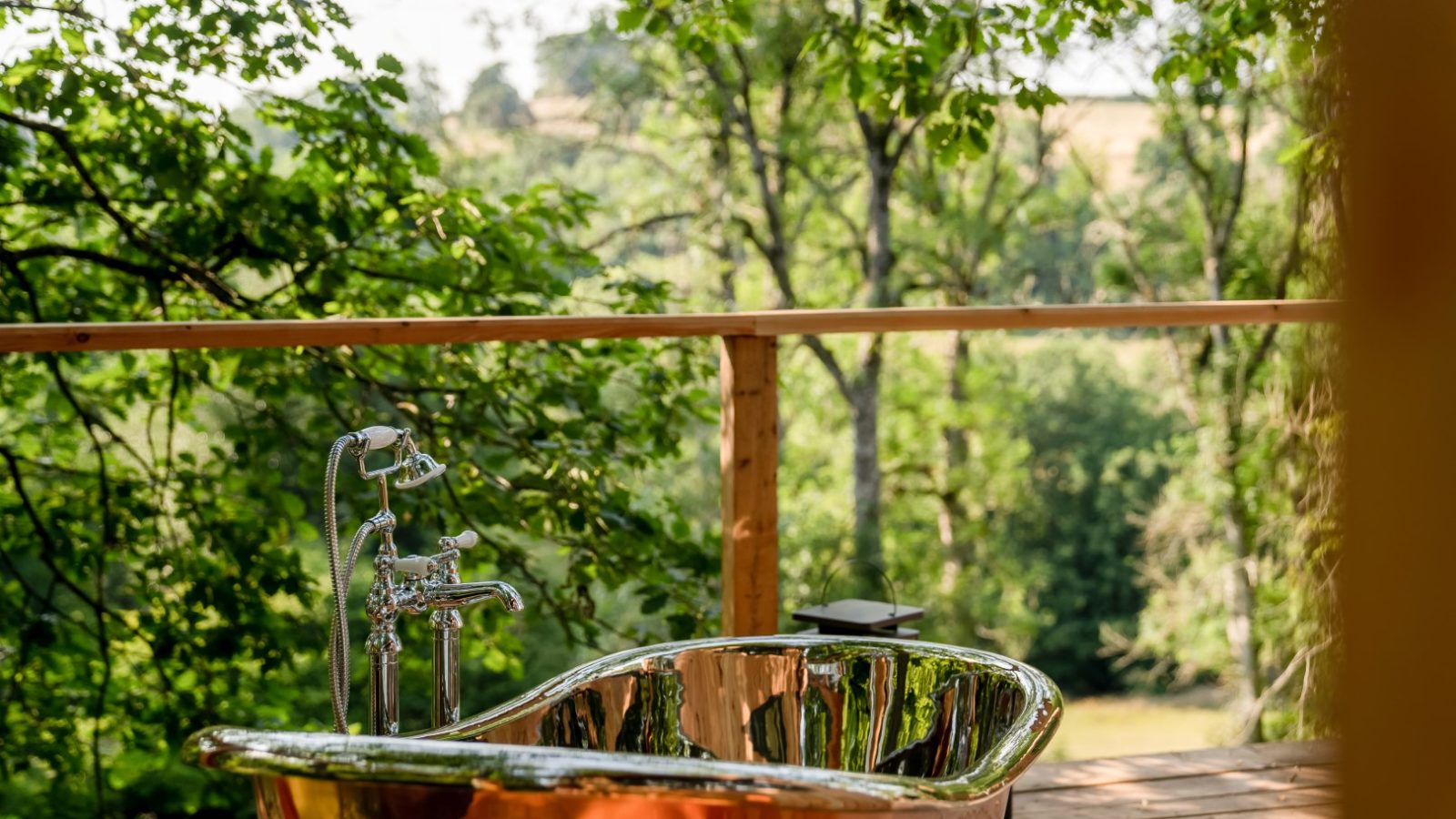 A copper bathtub on a wooden deck offers a serene escape at The Fold, surrounded by lush greenery and majestic trees, evoking a luxurious treehouse vibe.