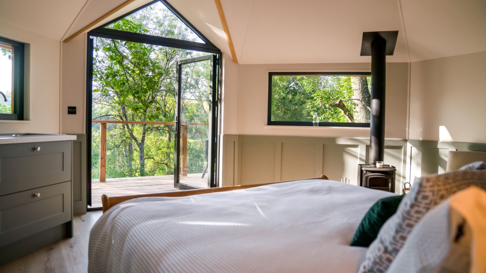 The bedroom, reminiscent of a cozy treehouse, boasts a large open window overlooking trees. It features a bed, a small wood stove, and light green walls that harmonize with nature's hues.