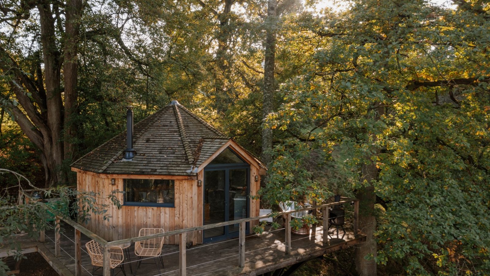 Nestled in the heart of a lush forest, The Fold's wooden treehouse boasts a conical roof and expansive windows. Surrounded by towering trees, its charming balcony with two chairs offers the perfect spot to soak in nature's tranquility.