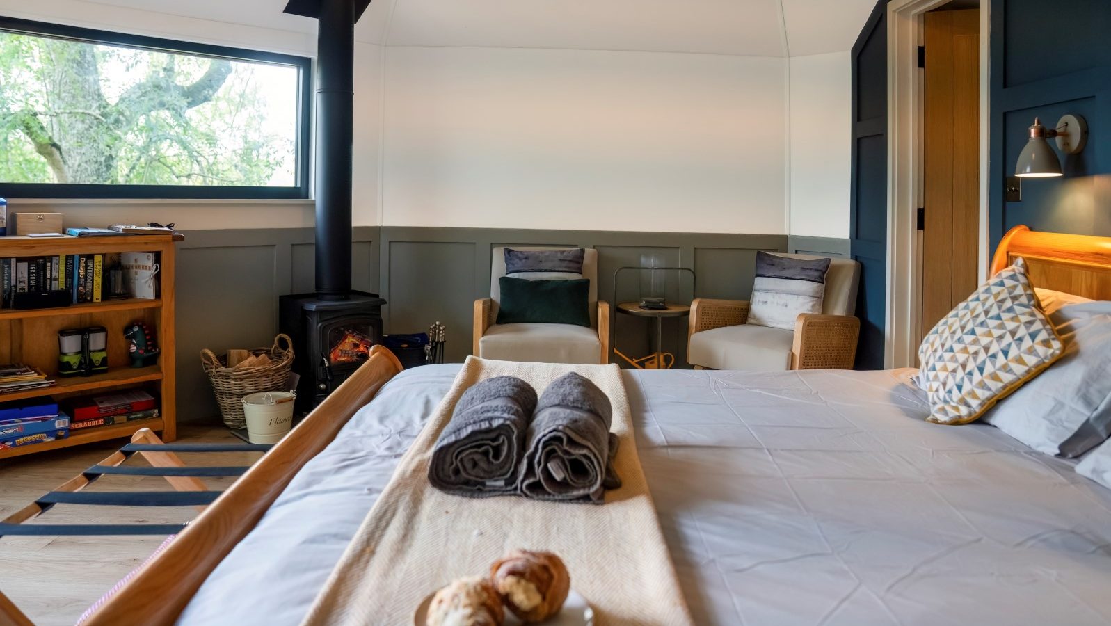 A cozy treehouse bedroom with a wood stove, bookshelves, two armchairs, and a bed adorned with neatly folded towels and pastries on a tray.