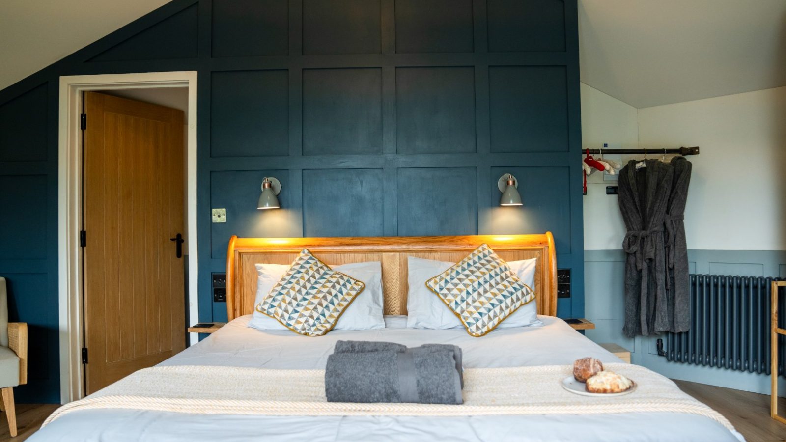 A cozy bedroom in The Fold, featuring a double bed with plush pillows, folded towels, and a calming blue accent wall. Two robes hang by the radiator, creating an inviting space that feels like your own personal treehouse retreat.