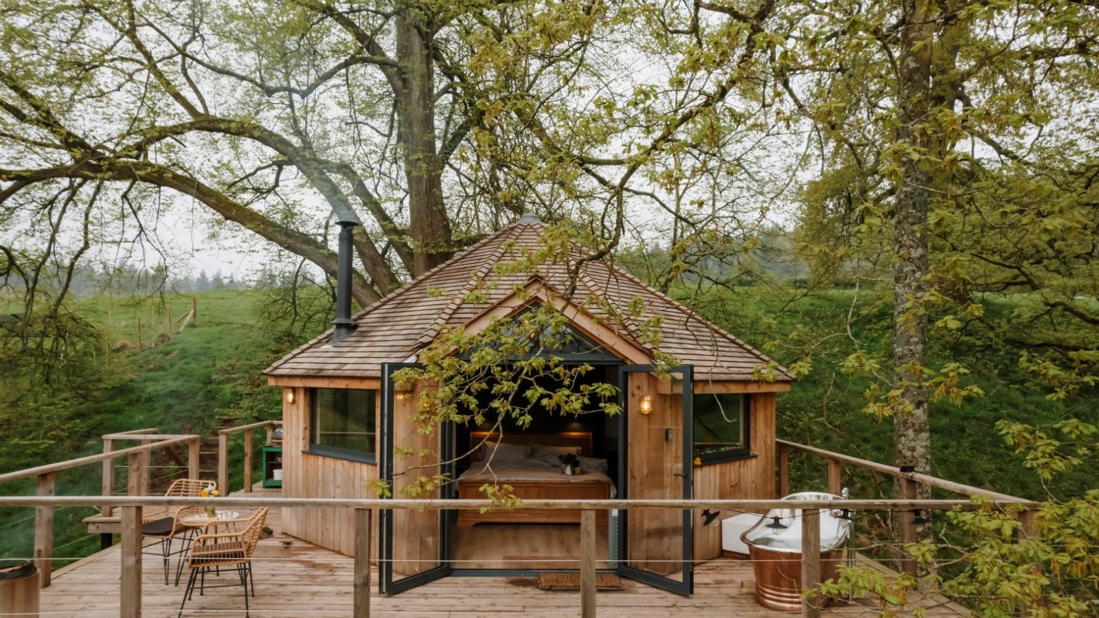 The Fold's wooden treehouse features open glass doors and is nestled among leafy trees, perched on a raised deck complete with outdoor seating and a bathtub.