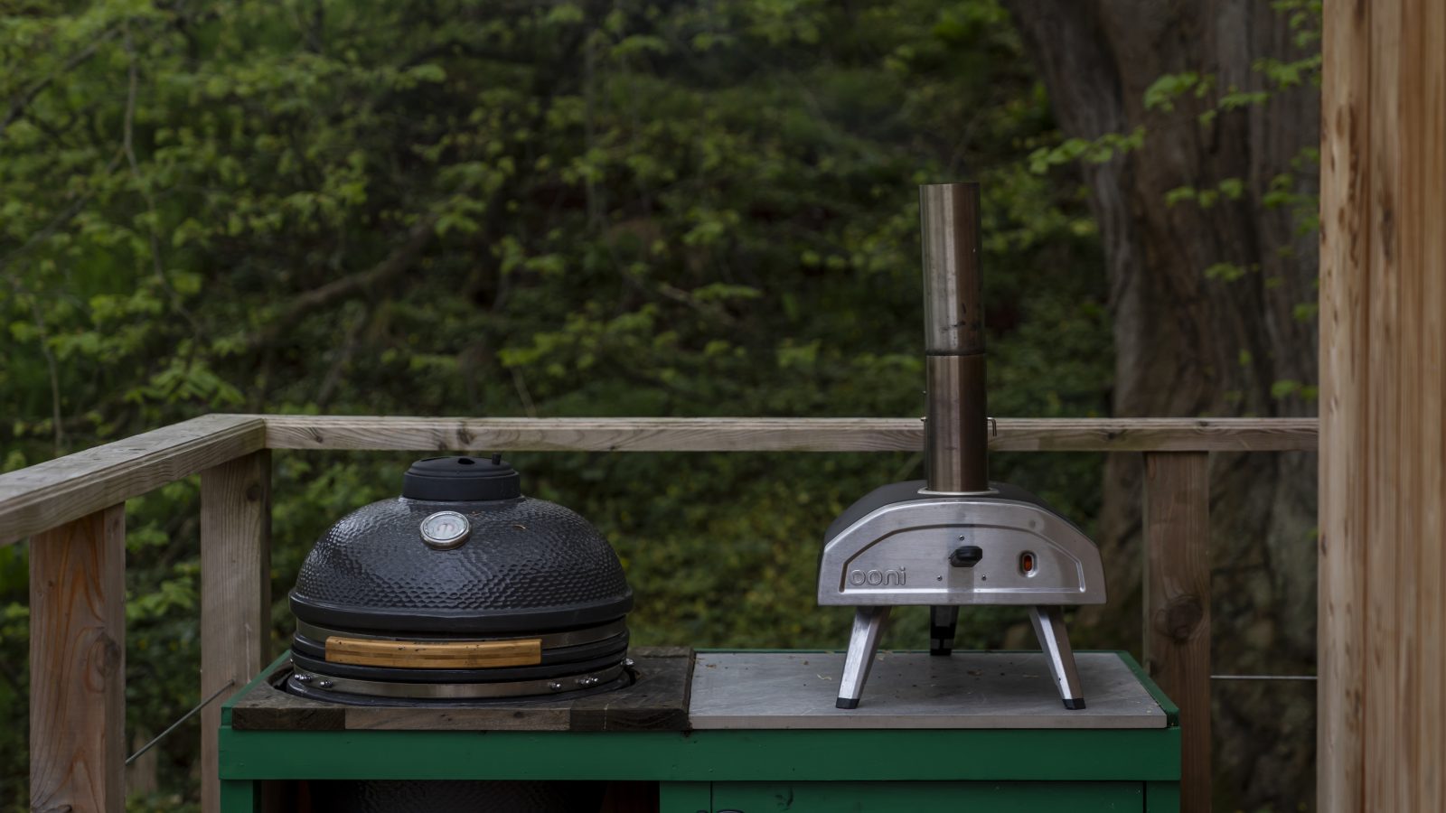 Nestled beside a treehouse, the outdoor setup features a green cabinet cradling a charcoal grill and a compact metal pizza oven, all embraced by lush greenery, reminiscent of The Fold's seamless connection with nature.