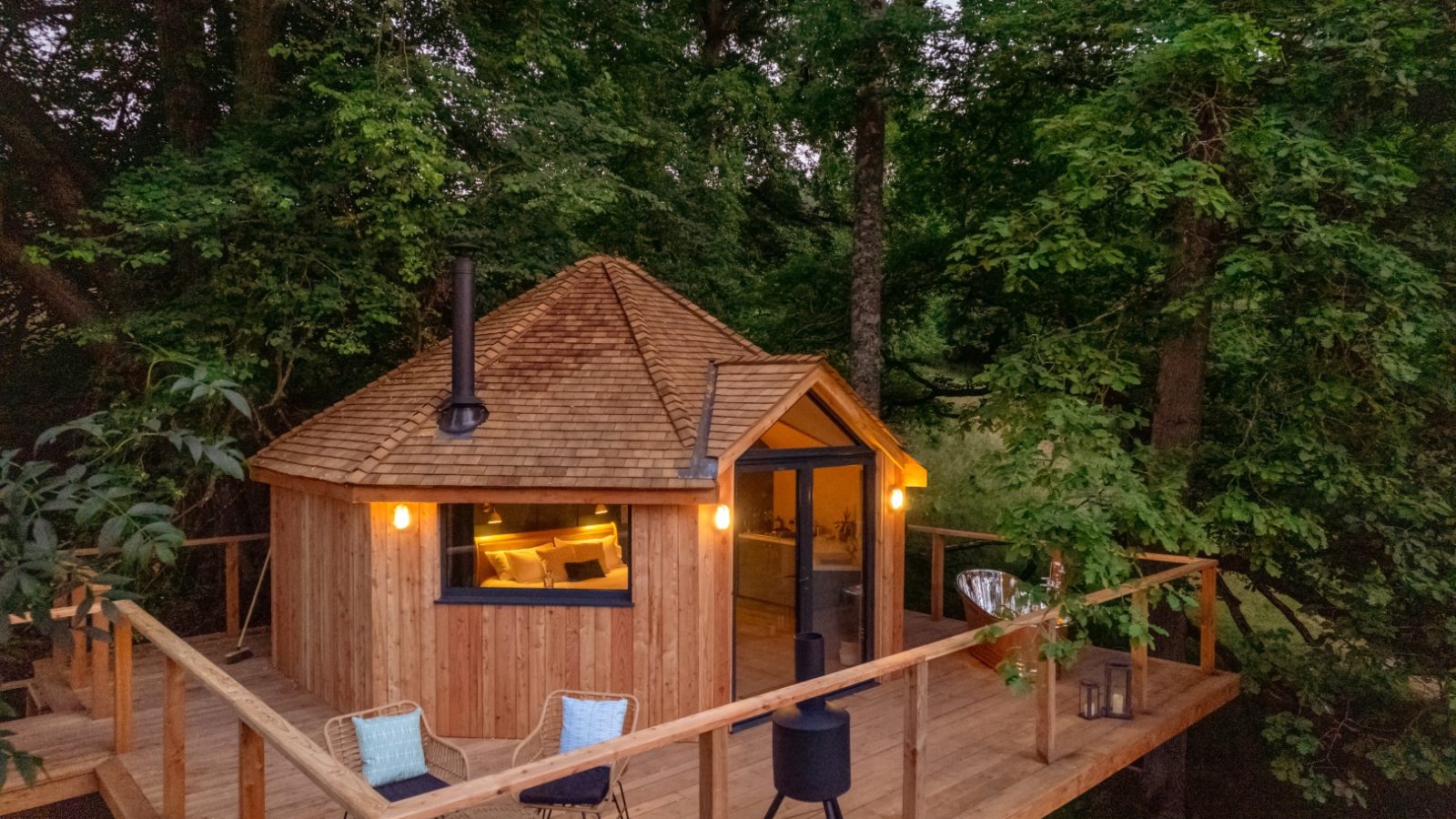 A charming treehouse nestled among trees, featuring a wooden deck with a cozy seating area. Warm lighting spills from The Fold within the cabin, creating an inviting ambiance inside and out.