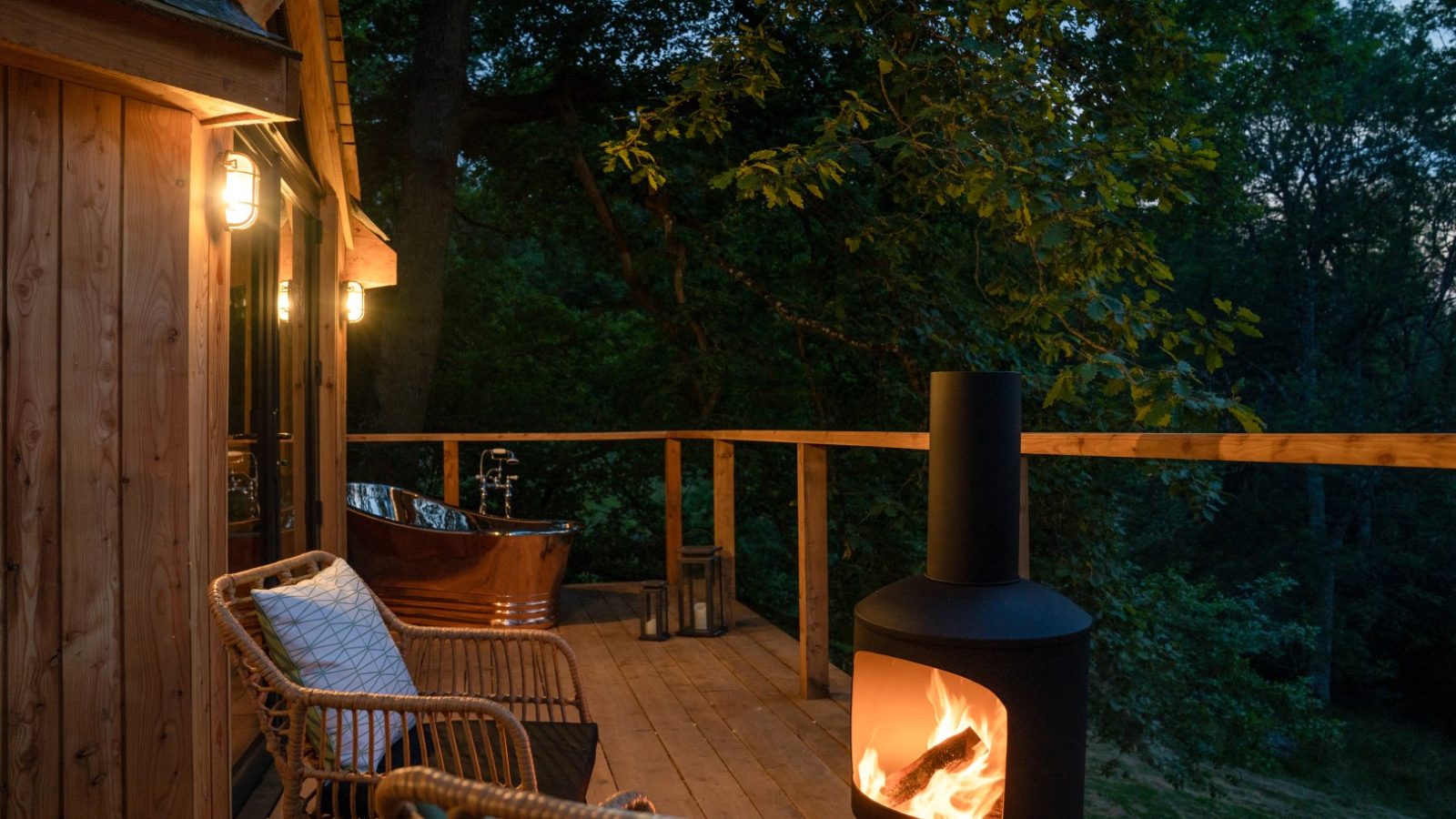 A rustic wooden deck outside The Fold features wicker chairs, a lit outdoor fireplace, and a copper bathtub, all enveloped by the serene surroundings of trees at dusk, creating the perfect treehouse vibe.