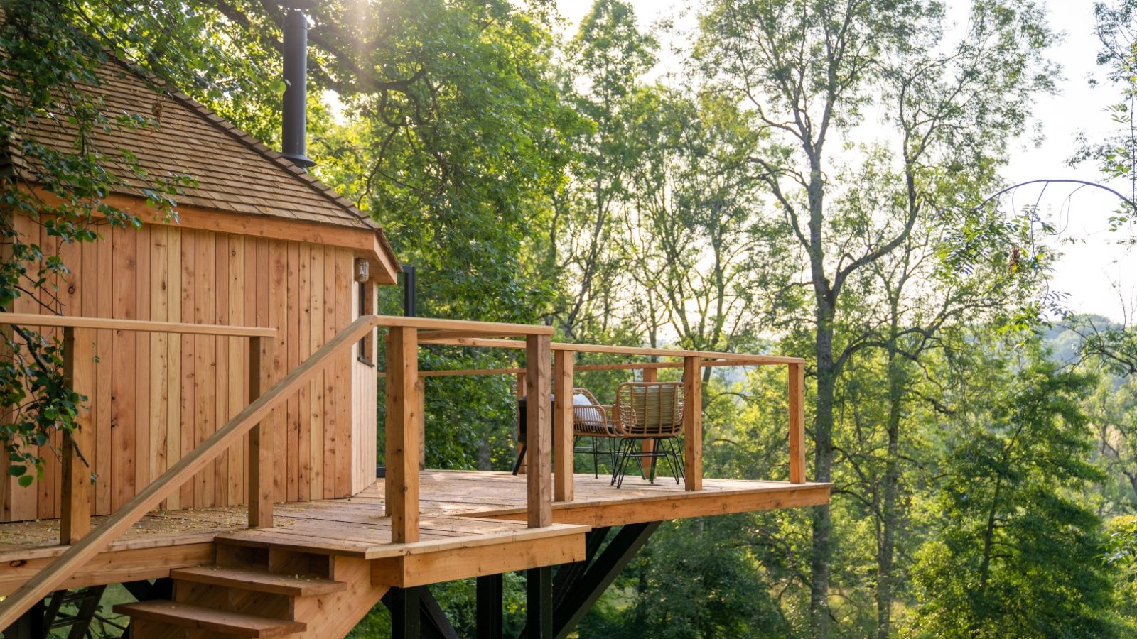 Nestled high above the ground, The Fold is a charming wooden treehouse with a deck and chairs, perfectly surrounded by lush green trees basking in sunlight.