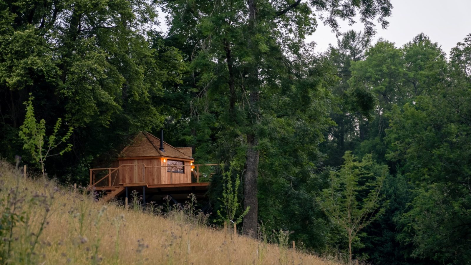 A small wooden cabin, reminiscent of a cozy treehouse, with lit windows is nestled among trees on a grassy hillside.