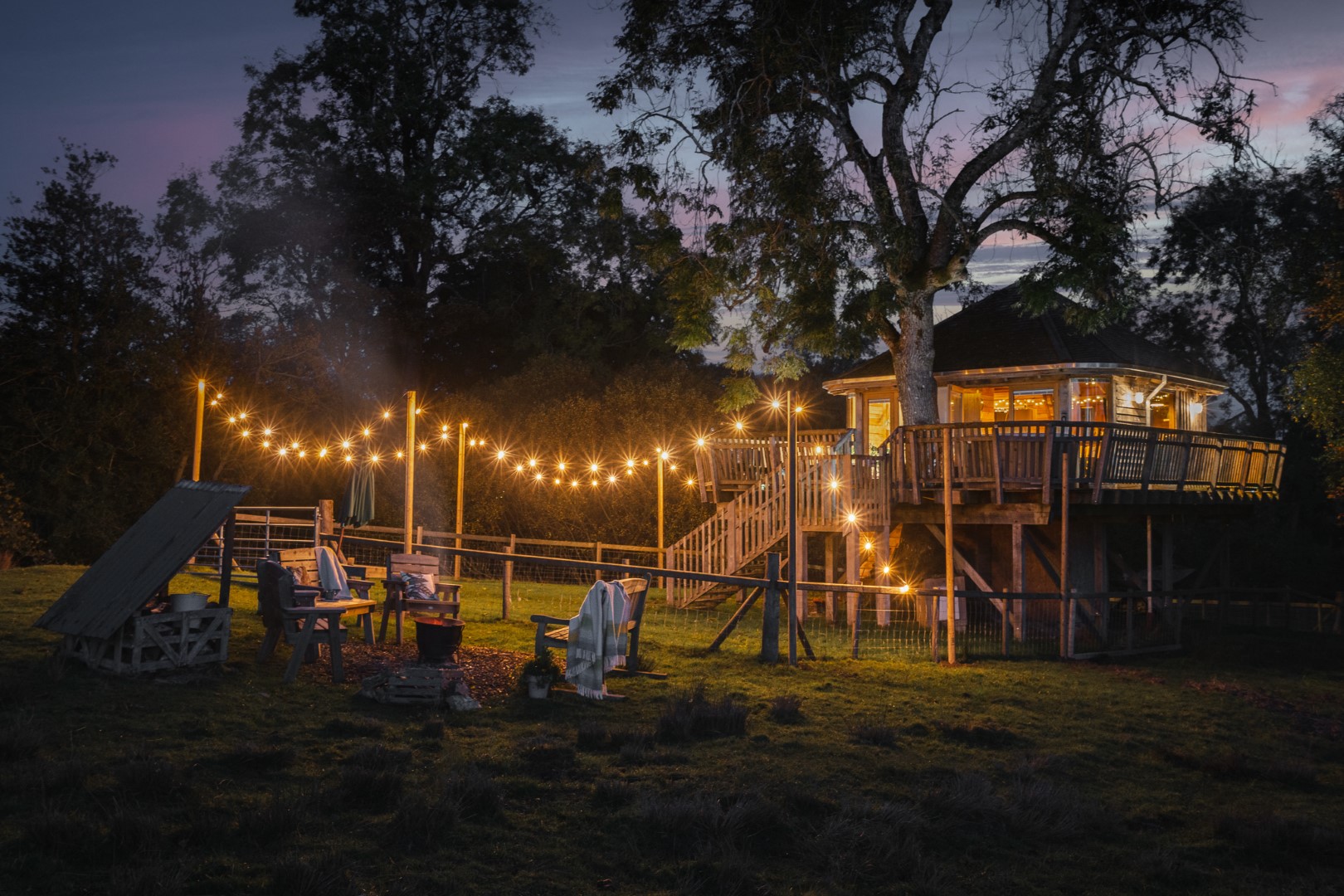 A treehouse, perfect for a dreamy giveaway, glows with string lights at dusk, offering a cozy outdoor seating area by the fire pit.