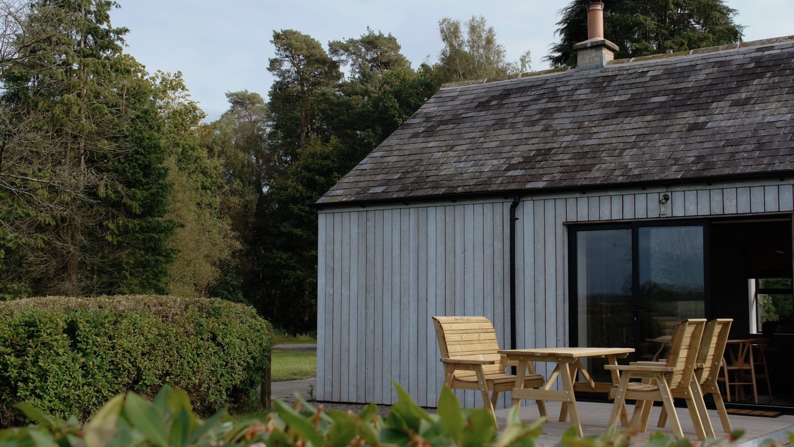 Nestled amidst lush greenery and trees, Beckside Cottage is a charming wooden cabin featuring a sloped roof and a cozy outdoor seating area.