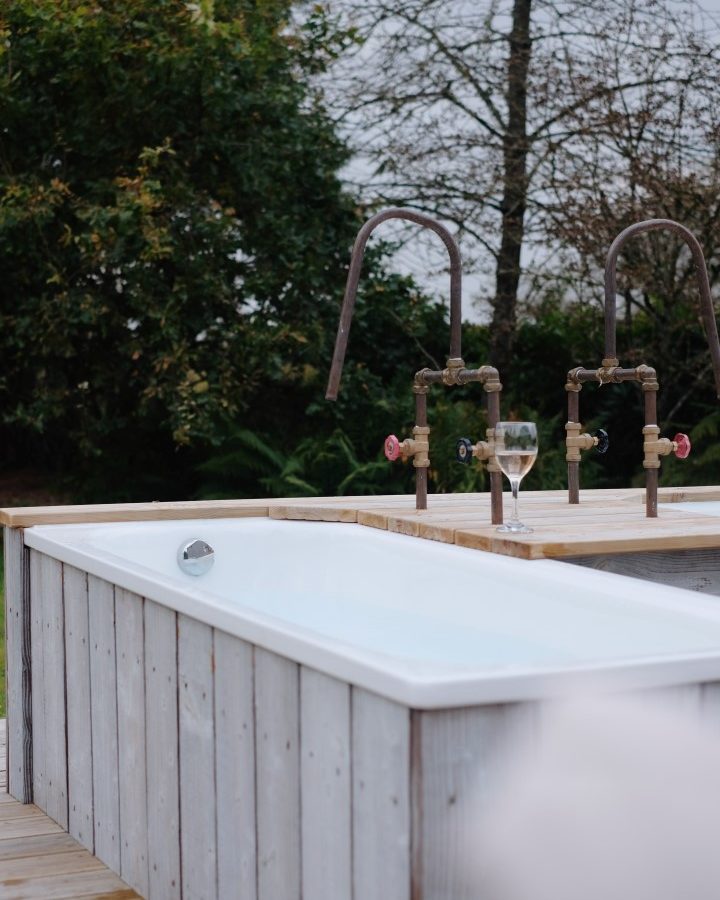 Nestled at Beckside Cottage, an outdoor wooden hot tub with water taps invites relaxation, complemented by a glass of white wine resting on the edge. Surrounded by trees, this tranquil setting promises a rejuvenating retreat in nature’s embrace.