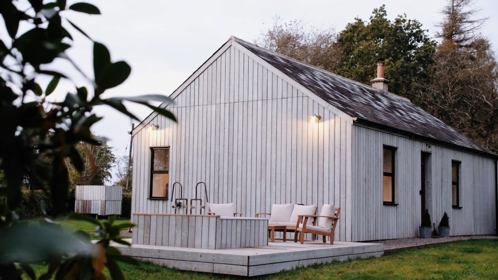 Nestled amidst lush greenery, Beckside Cottage is a charming, small white wooden retreat featuring an inviting outdoor deck with seating. The tranquil setting offers a perfect escape into nature.