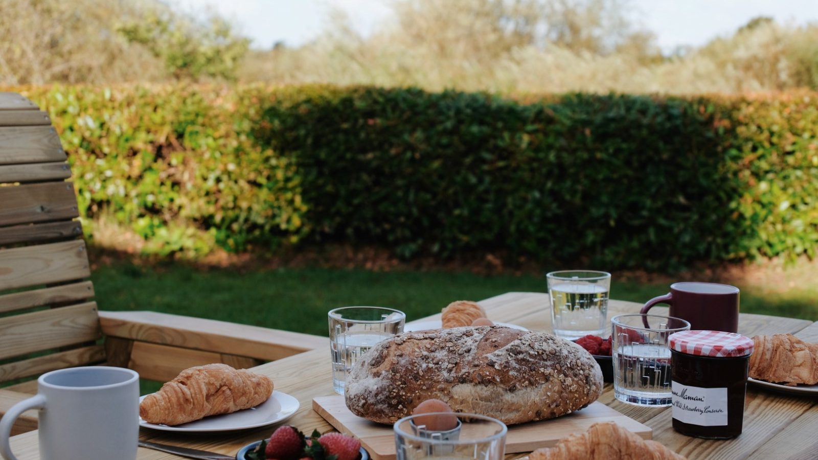 An outdoor table at Beckside Cottage is adorned with bread, croissants, strawberries, jars, and mugs. Greenery in the background thrives under a clear sky.