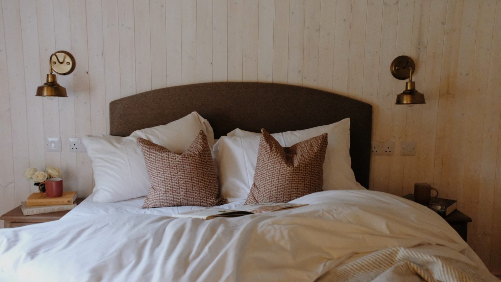 A neatly made bed with two brown pillows, white bedding, and a dark headboard graces Beckside Cottage. Wall lamps illuminate the wooden backdrop, adding to its cozy charm.