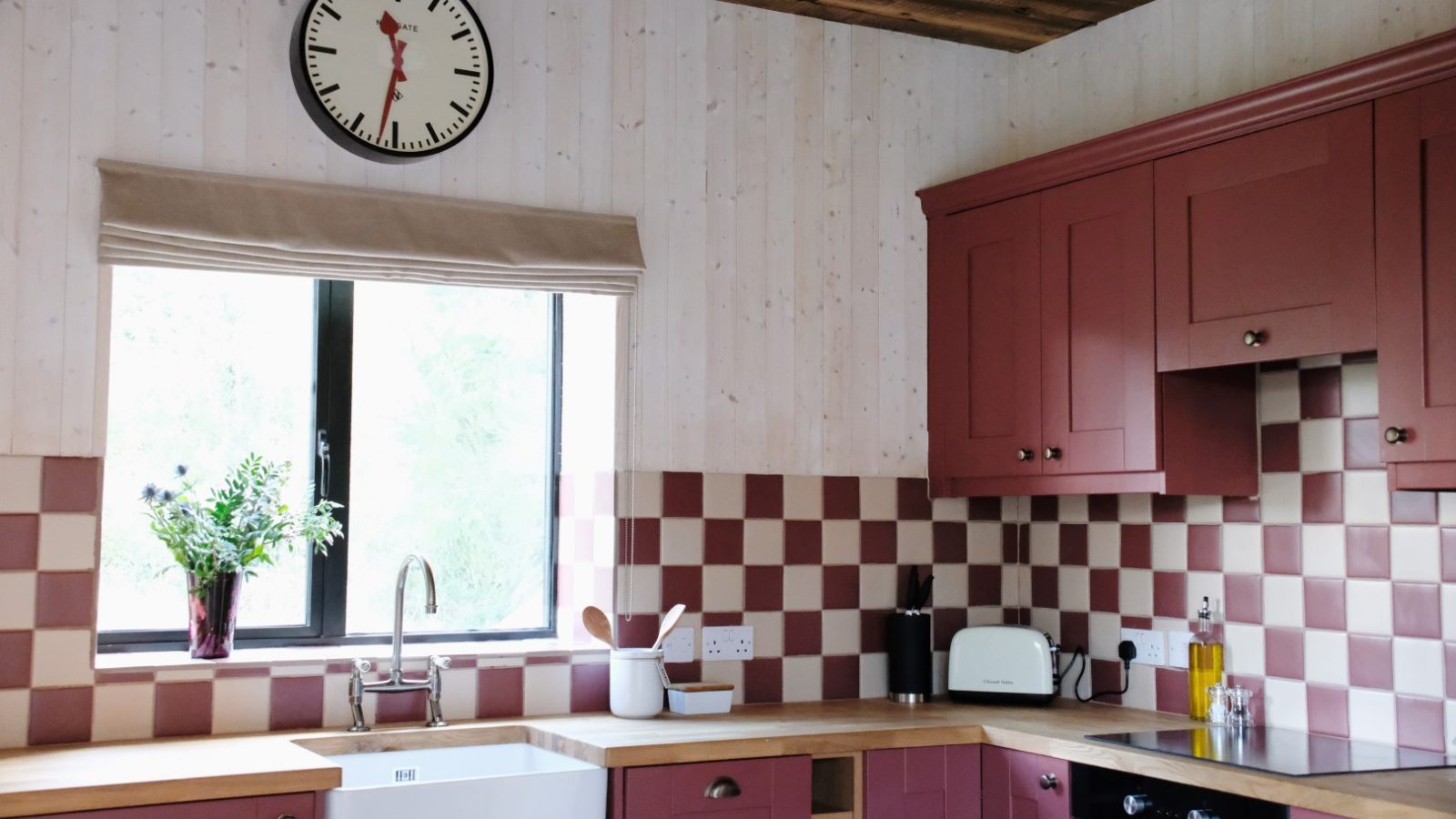 The cozy kitchen at Beckside Cottage features maroon cabinets, checkered tiles, a large clock, and a window with a potted plant on the counter.