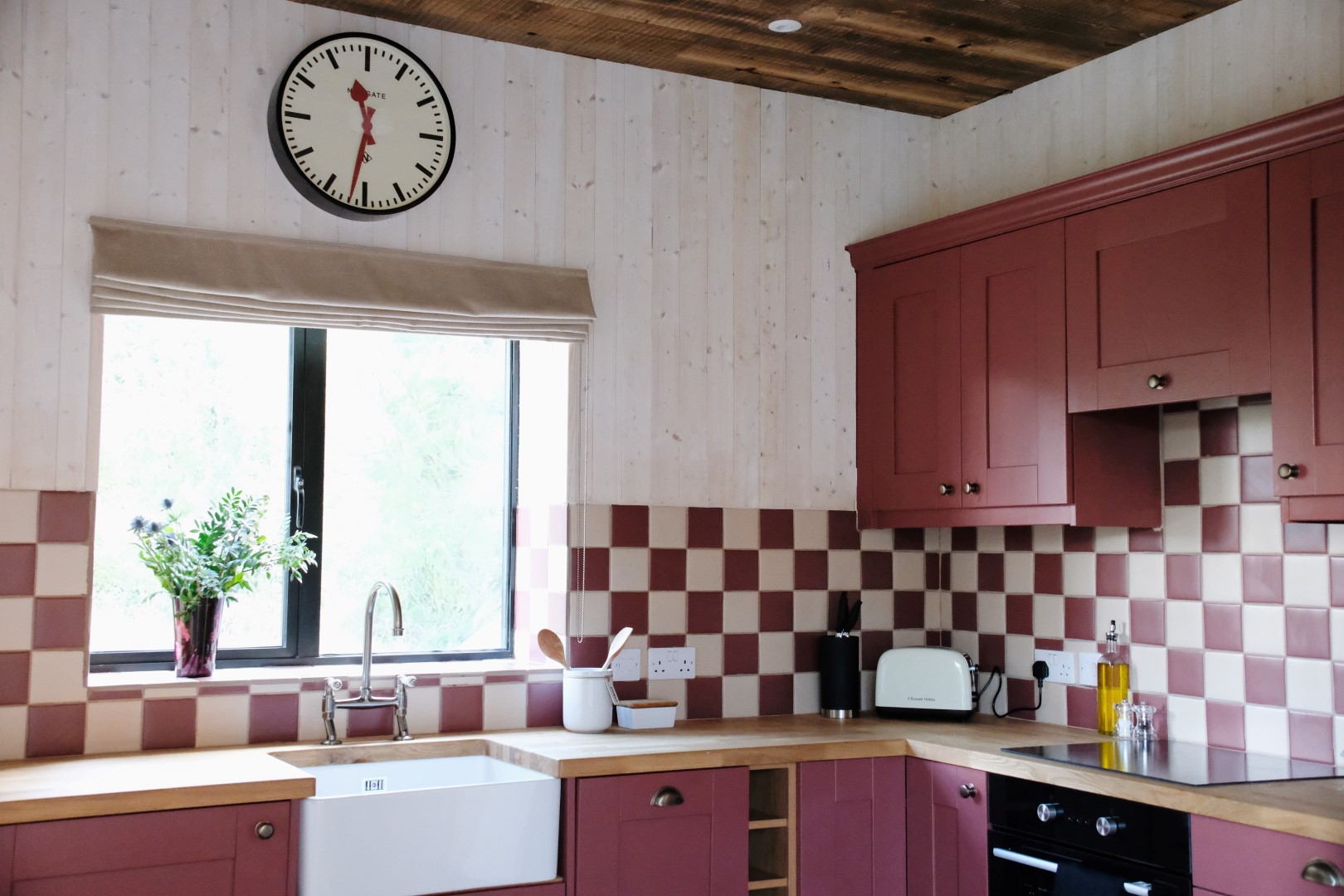 The cozy kitchen at Beckside Cottage features maroon cabinets, checkered tiles, a large clock, and a window with a potted plant on the counter.