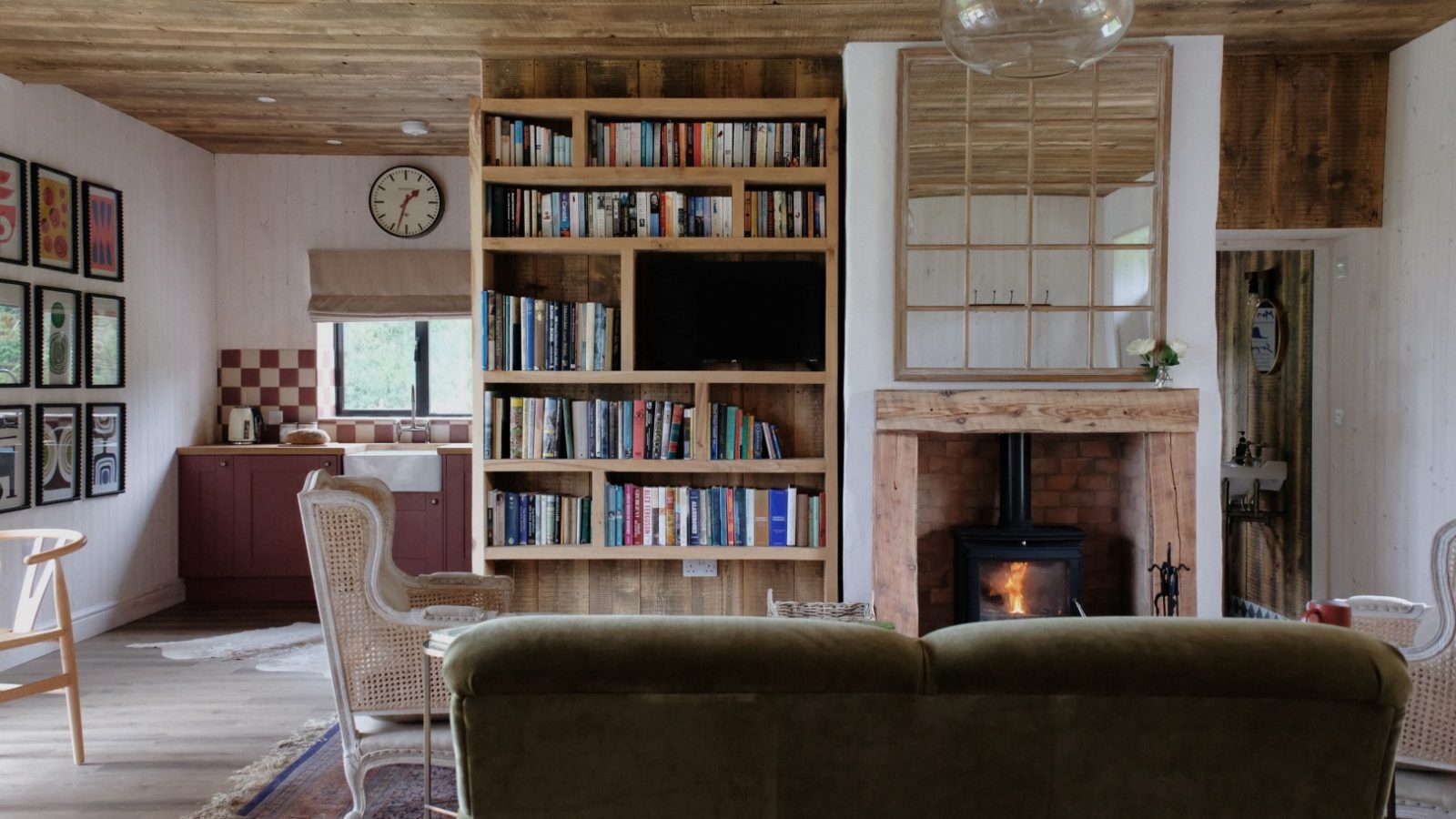 Cozy living room in Beckside Cottage with a bookshelf, armchairs, and a wood-burning stove. In the background, the kitchen adds to the charm. A clock on the wall reads 12:15.