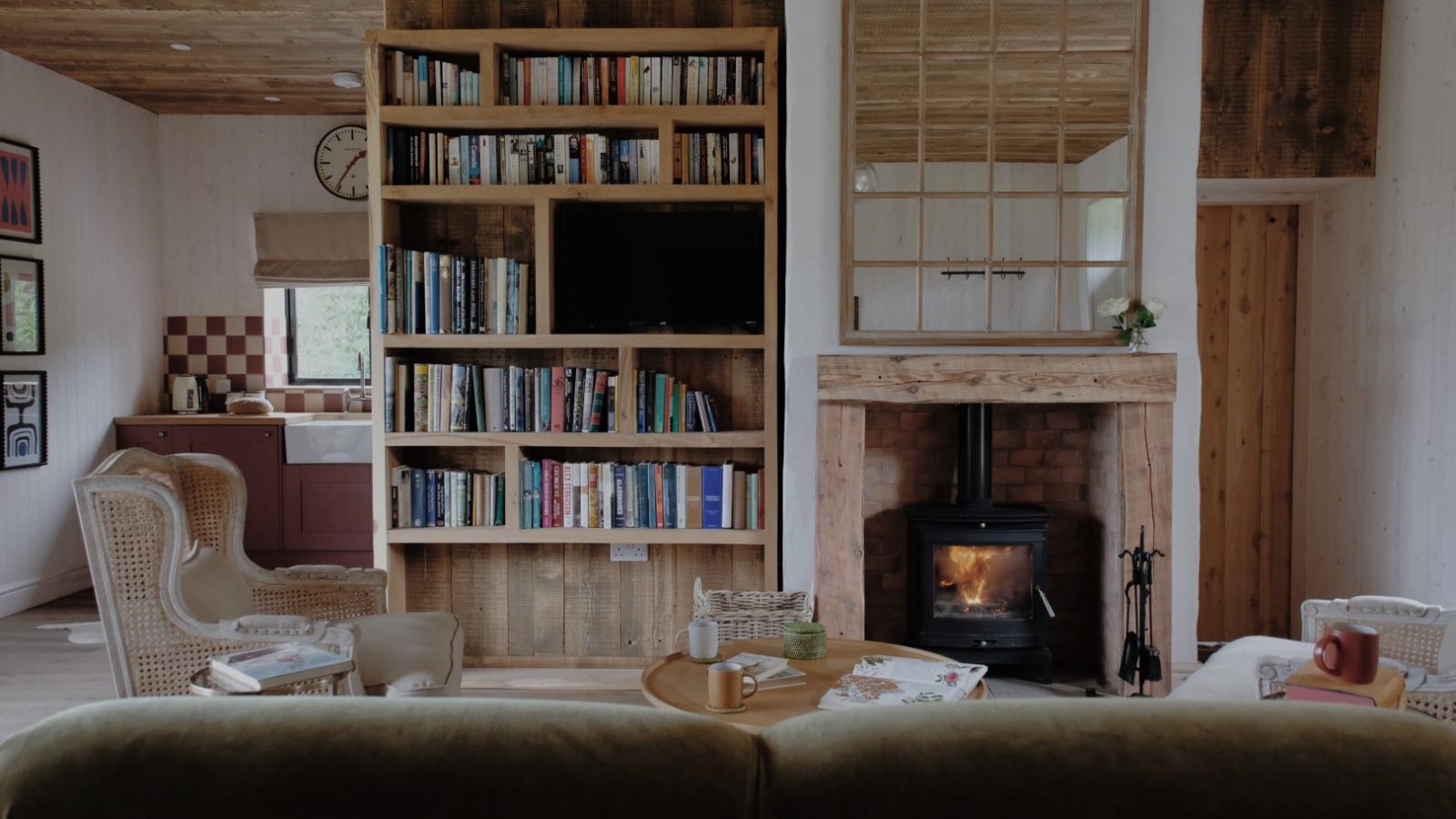 Cozy living room in Beckside Cottage, complete with a bookshelf, TV, wood-burning stove, and wicker chairs. Light streams in from windows above the sink.