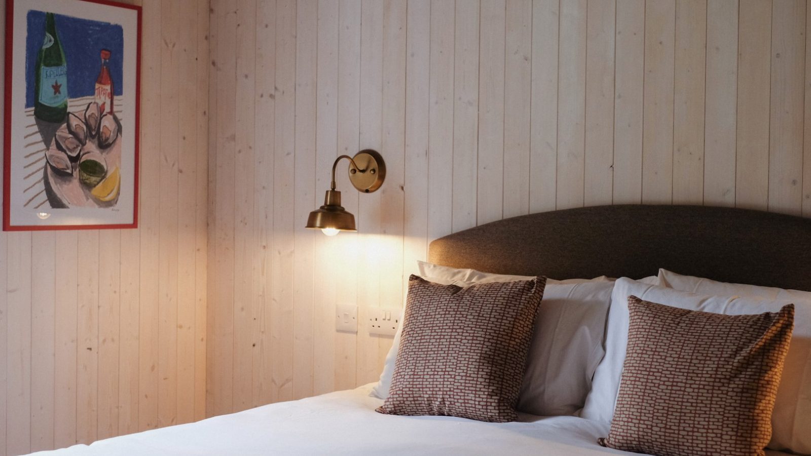 Cozy bedroom at Beckside Cottage with wood-paneled walls, a bed adorned with brown pillows, a wall-mounted lamp, and a framed still-life painting.