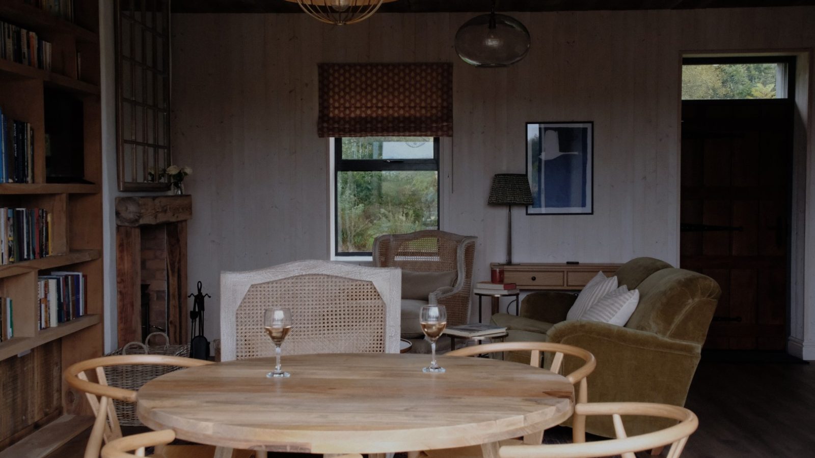 The cozy living room at Beckside Cottage features wooden furniture, bookshelves, a fireplace, and two wine glasses elegantly placed on the round table.