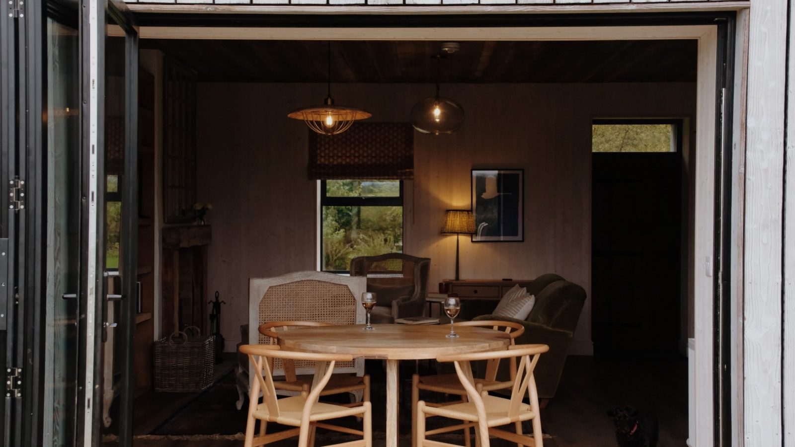 Open wooden doors reveal a cozy room at Beckside Cottage, complete with a round table, chairs, a comfy couch, lamps, and a window offering a serene view of lush greenery.