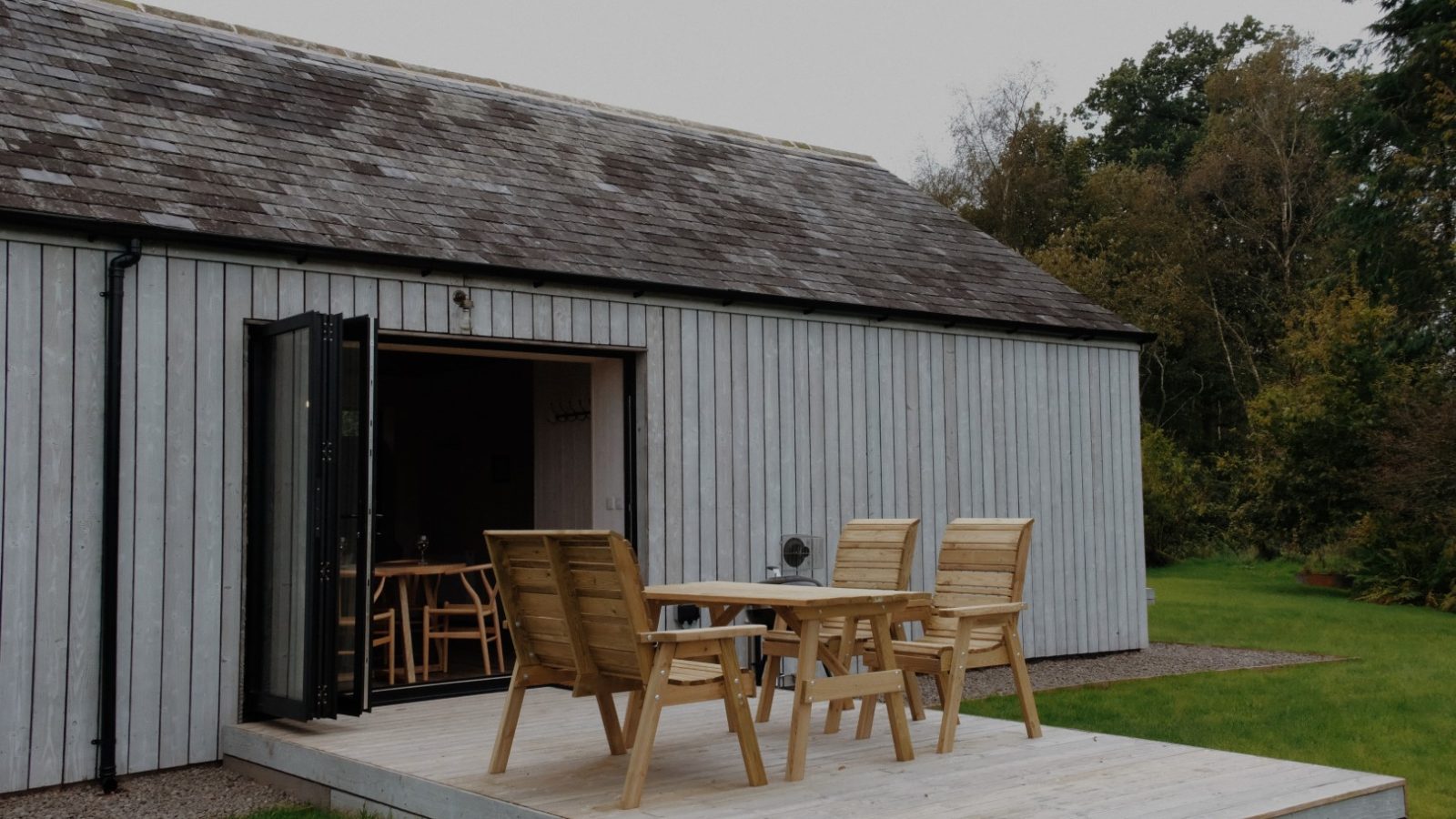 Wooden chairs and a table grace the small deck of Beckside Cottage, outside a gray wooden building with open folding doors inviting you inside.