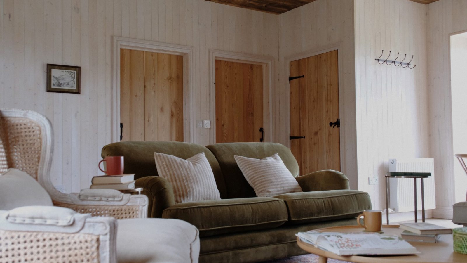 The cozy living room at Beckside Cottage features a green sofa, white armchairs, and a wooden ceiling. Coffee mugs and books are thoughtfully placed on the table, inviting you to unwind.