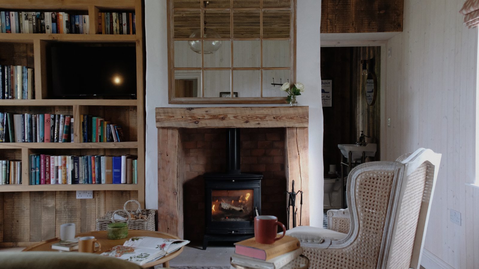 The cozy living room at Beckside Cottage features a wood-burning stove, a comfy armchair, bookshelves, and a coffee table adorned with books and a steaming mug.