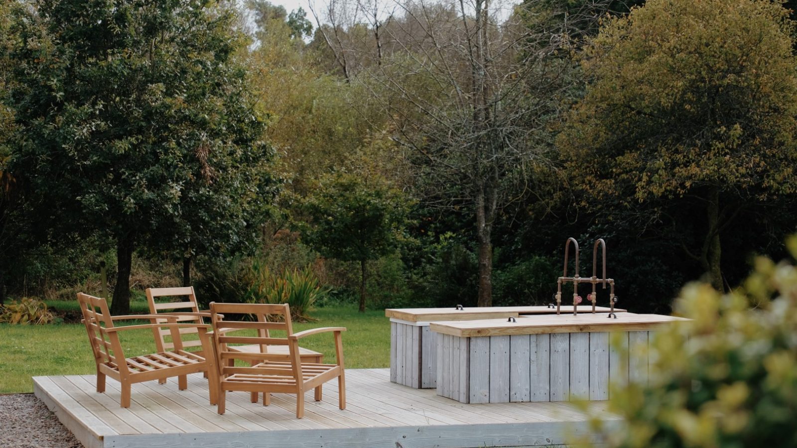 The wooden deck at Beckside Cottage features two chairs and a water basin, nestled in a serene garden setting surrounded by lush trees and greenery.