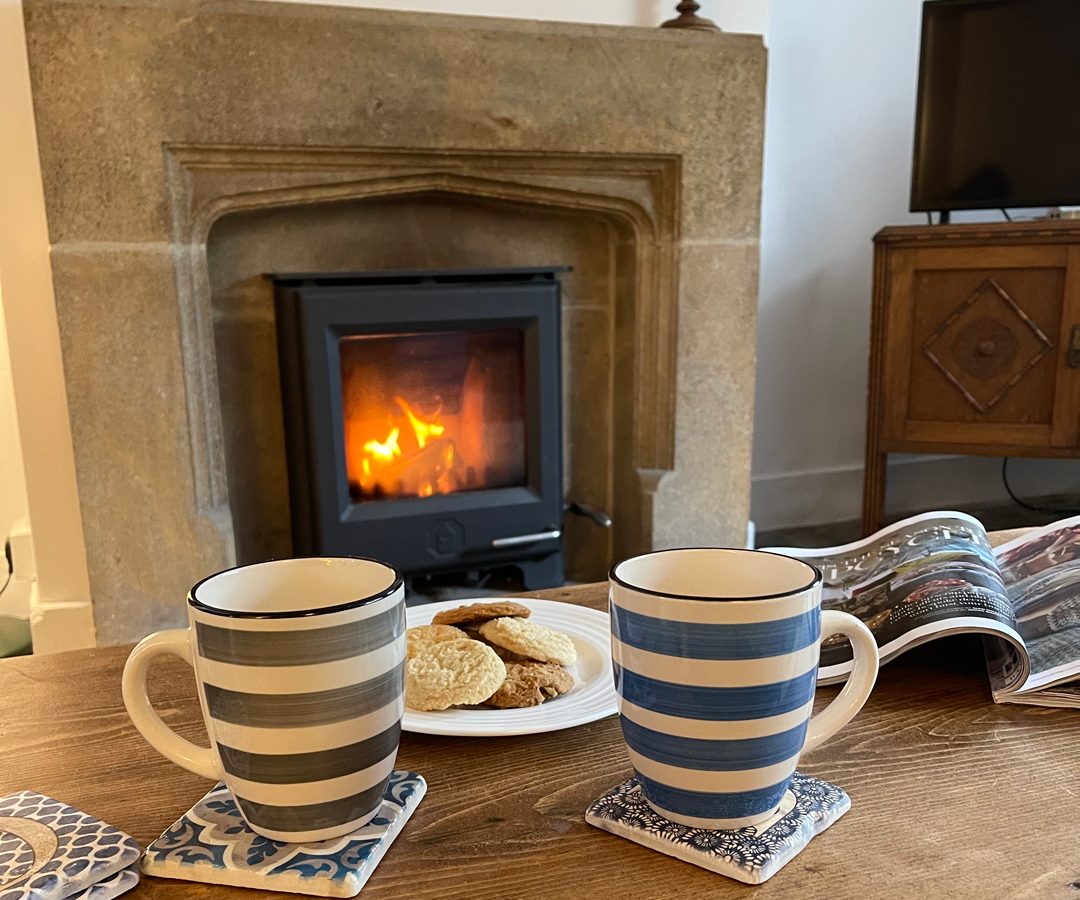 Two striped mugs and cookies on a table at Blackbird Cottage, with a fireplace glowing, a magazine lies open nearby.