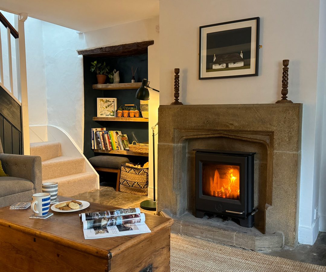 Cozy Blackbird Cottage living room with a lit fireplace, bookshelf, armchair, and coffee table filled with mugs and snacks.