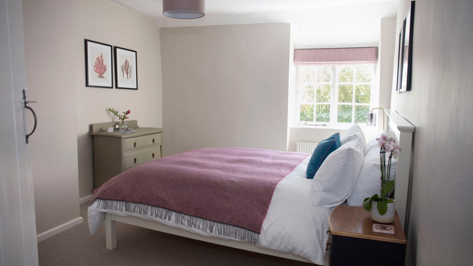 A small bedroom at Blackbird Cottage with a double bed, pink blanket, dresser, side table, pictures, and natural light.