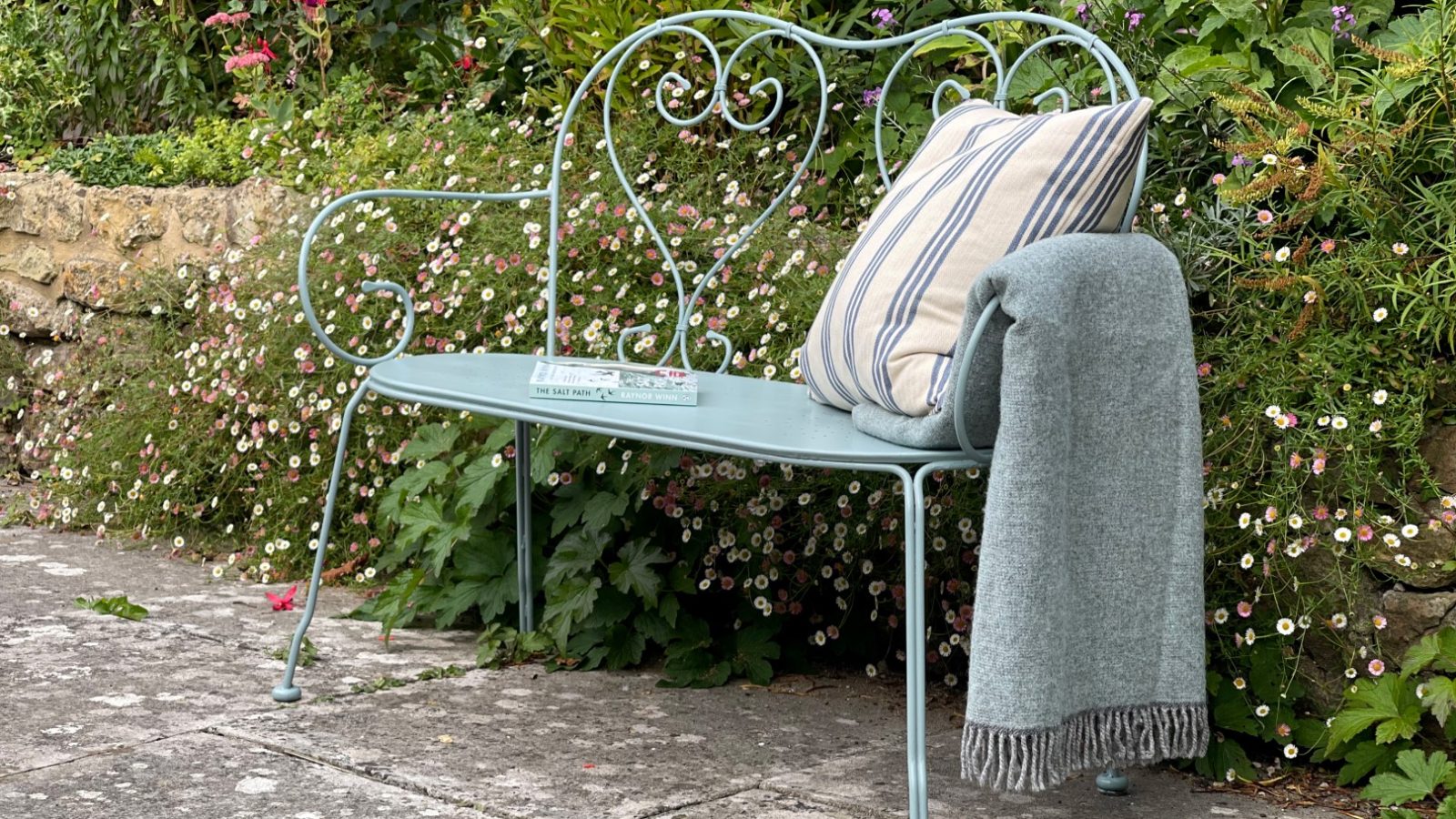 A decorative metal bench with a pillow and blanket sits on Blackbird Cottage's stone patio, surrounded by lush plants and flowers.