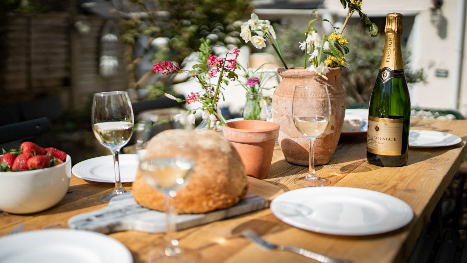 Enjoy a casual dining ambiance at Blackbird Cottage with a charming outdoor table set, wine, bread, and fresh strawberries.