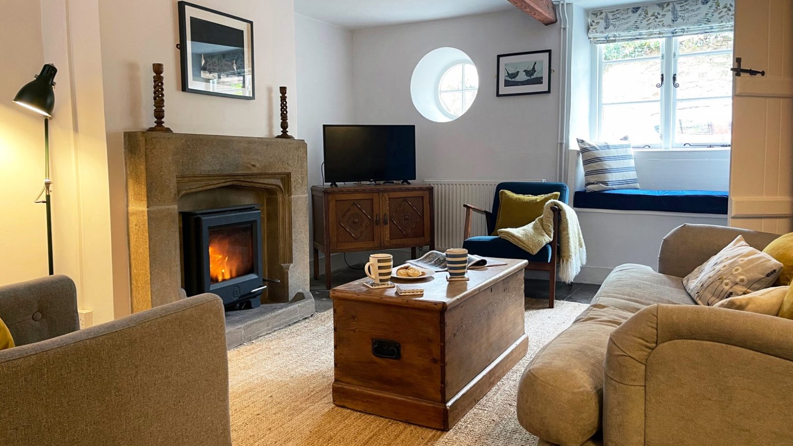 Cozy living room at Blackbird Cottage: a lit fireplace, TV, armchairs, wooden coffee table with cups, and window seat with cushions.