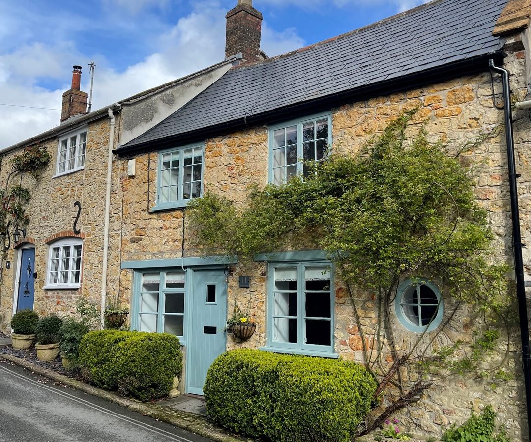 Blackbird Cottage features blue doors and windows, surrounded by green shrubs and climbing plants, on a narrow street.