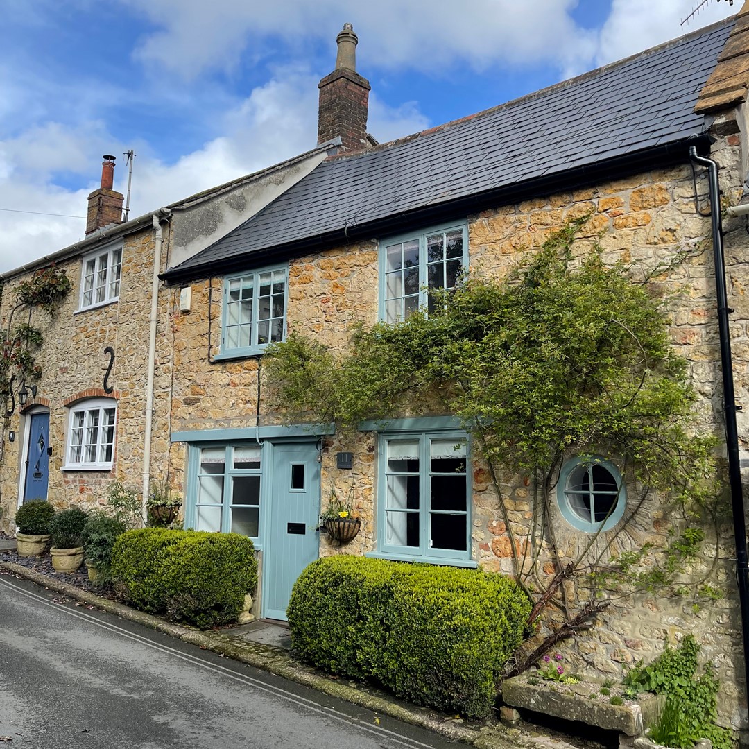 Blackbird Cottage features blue doors and windows, surrounded by green shrubs and climbing plants, on a narrow street.