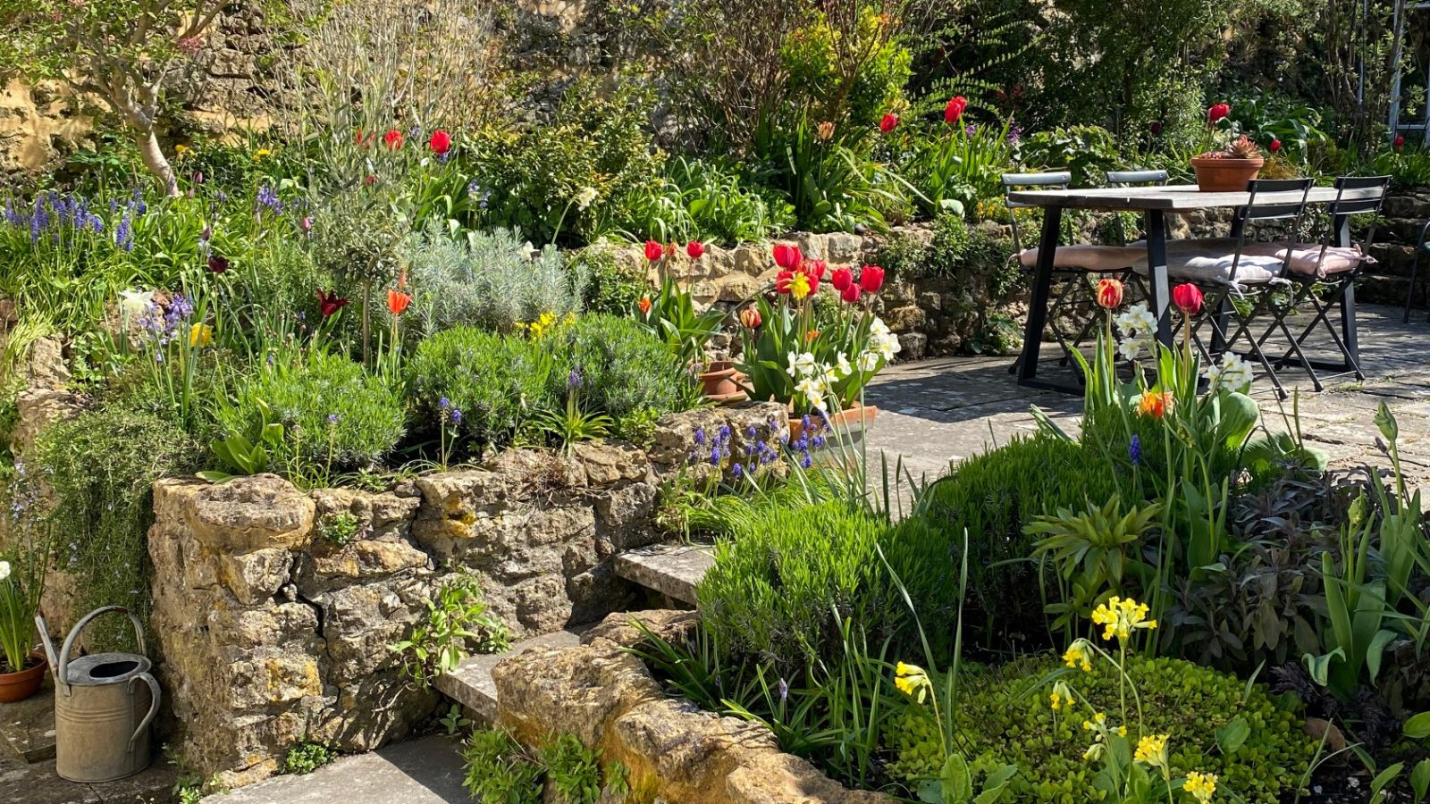 A lush garden at Blackbird Cottage with tulips, plants, a stone wall, and an outdoor table set on a sunny day.