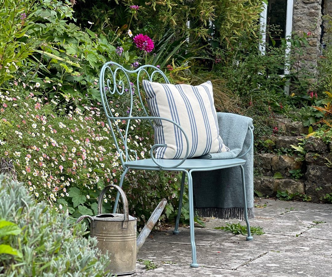 A metal garden bench at Blackbird Cottage, with a striped pillow, blanket, and a watering can on the stone path nearby.