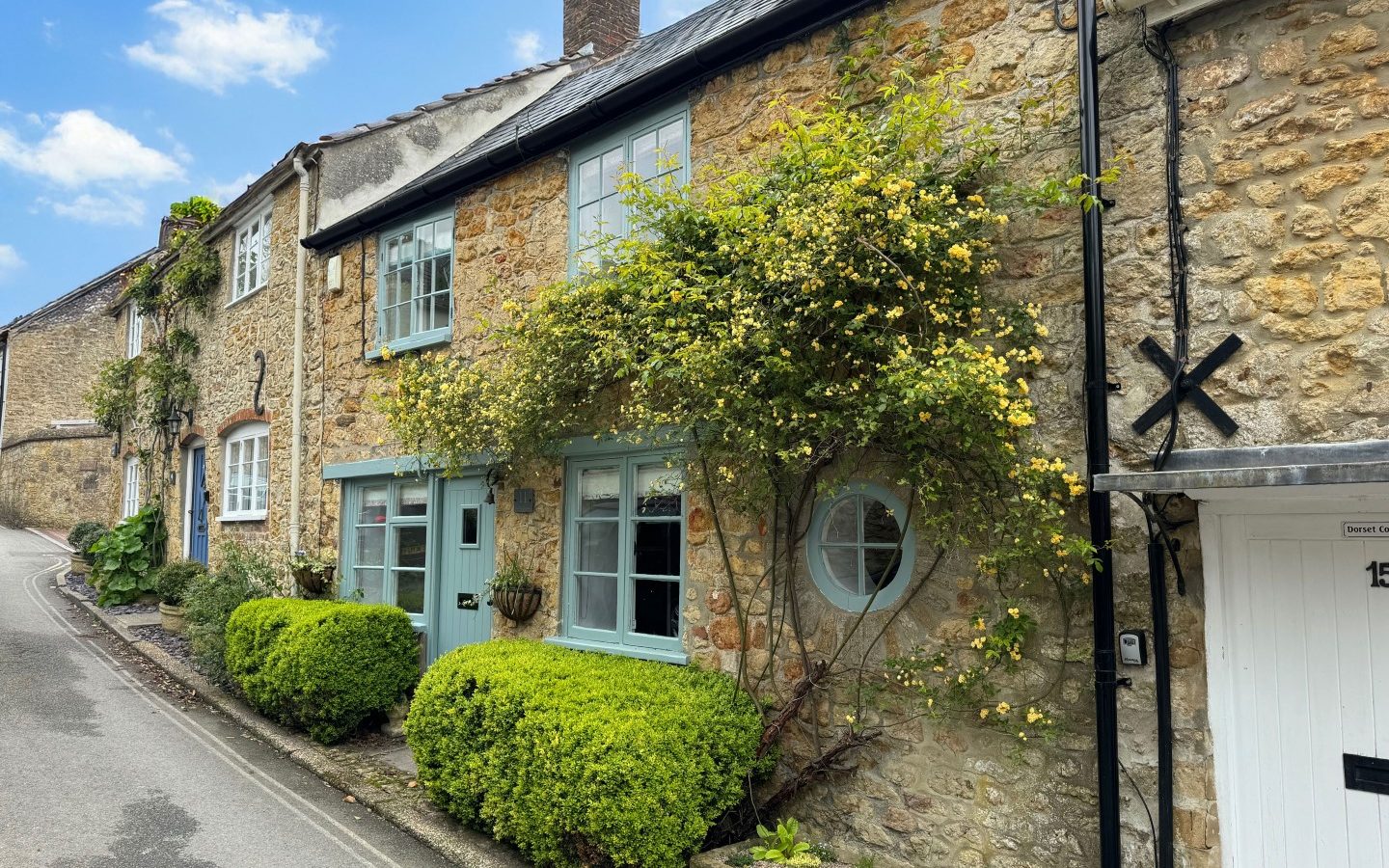 Blackbird Cottage: A stone retreat with blue window frames and climbing plants on a narrow street under a partly cloudy sky.