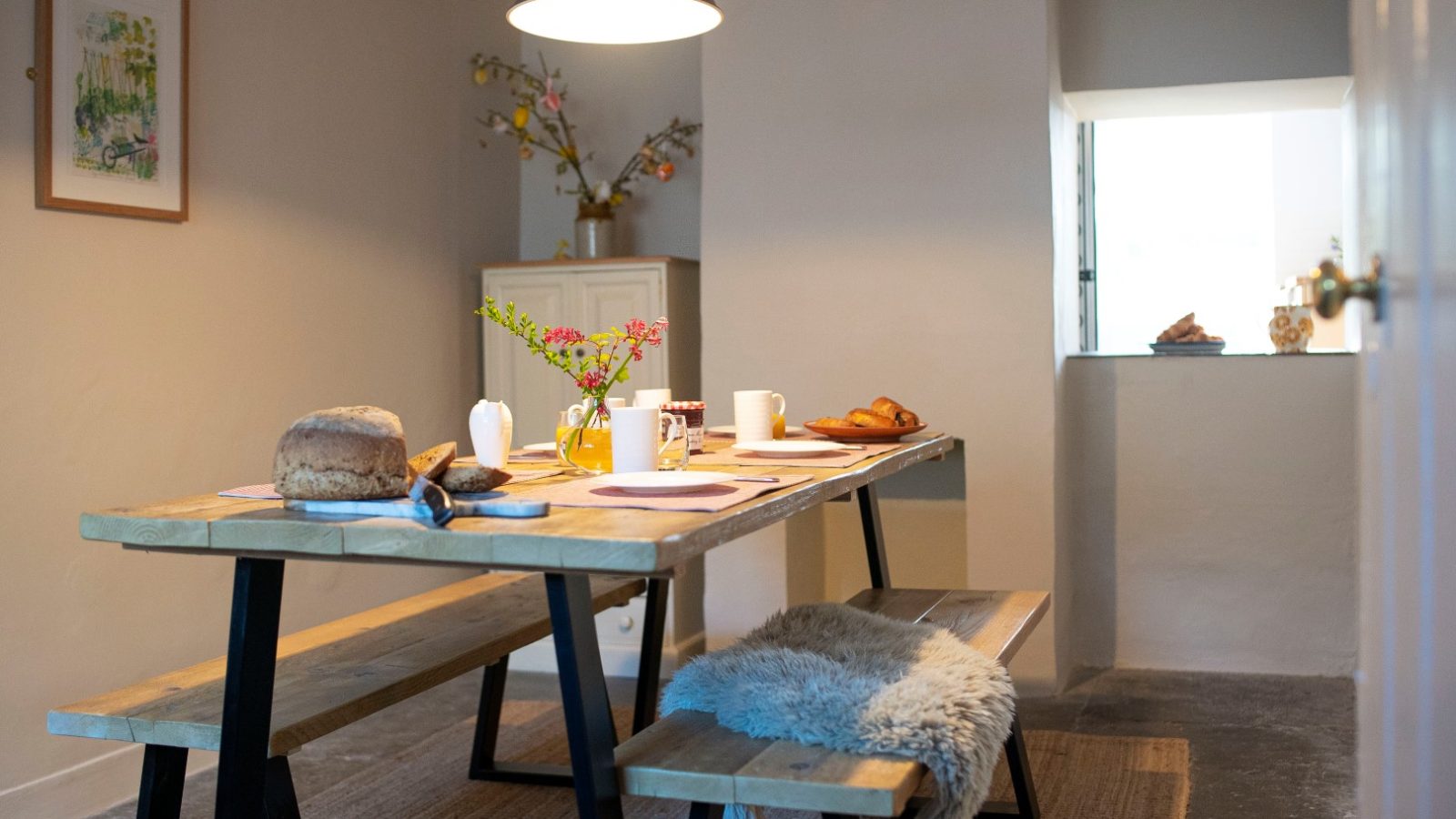 Rustic Blackbird Cottage dining area with a wooden table, bread, croissants, a plant, bench, and soft lamp lighting.